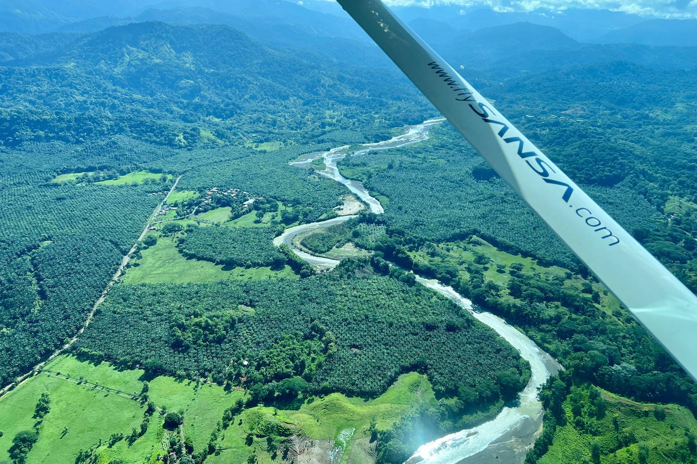 View from the window of a small hopper plane — the wing above a river winding through green mountains and palm plantations