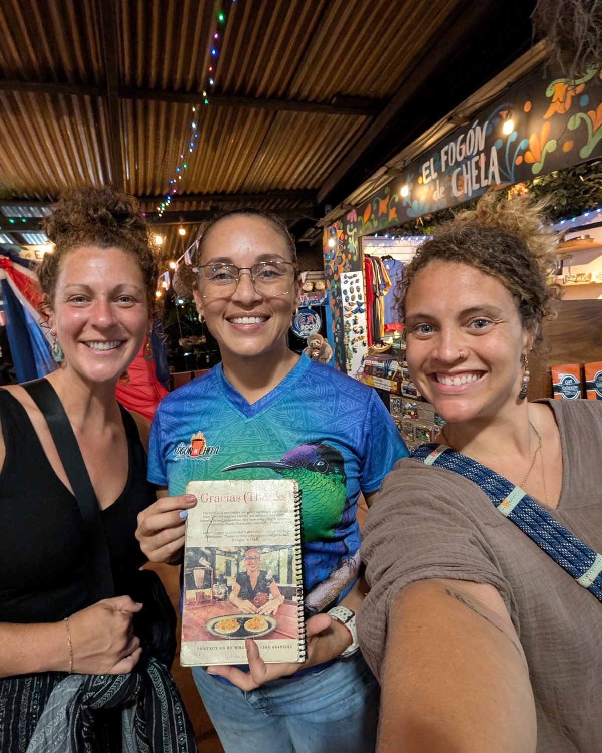 Tasha and a friend either side of Chela, smiling in her open-air kitchen under string lights, holding her handwritten recipe book