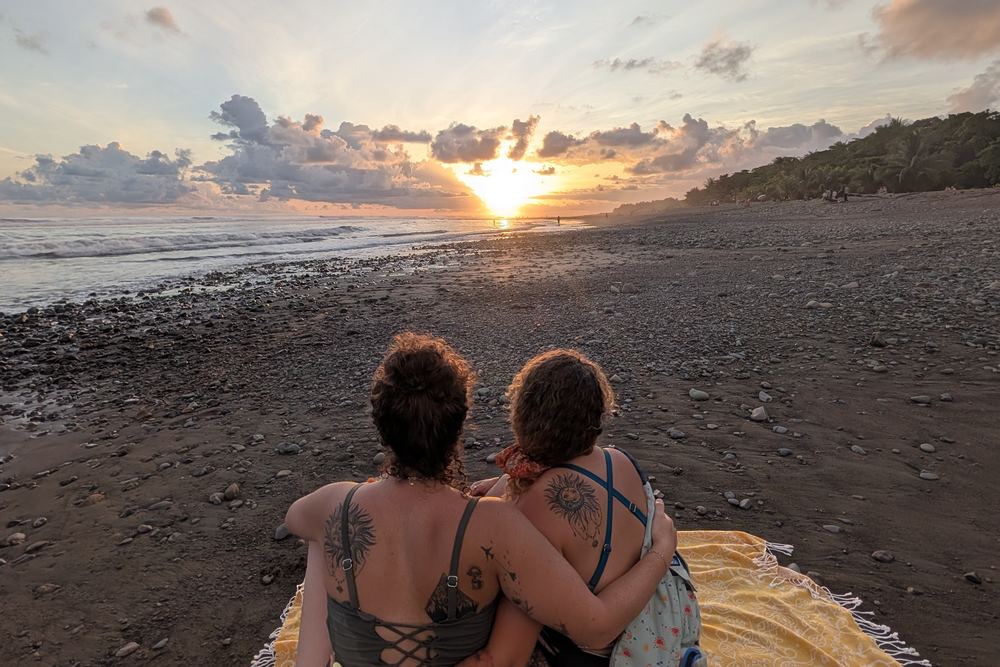 Two friends sitting close on a yellow blanket on Dominical Beach, watching the sun drop into the Pacific
