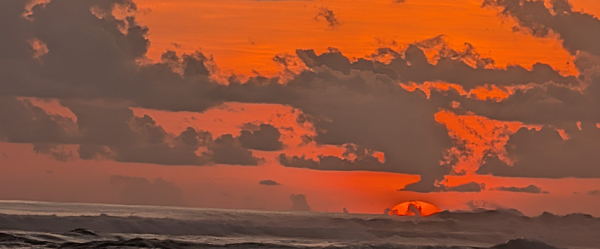 The sun dropping into the Pacific behind a bank of cloud, sky burning orange over the surf
