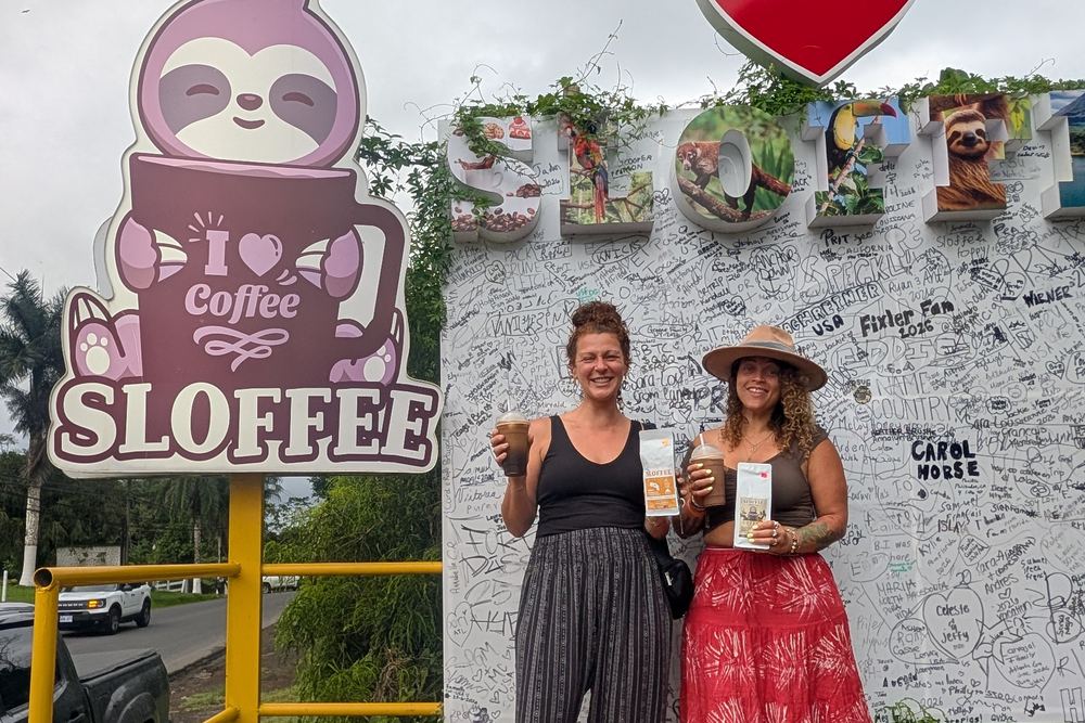 Tasha and a friend with iced coffees under the Sloffee sign, in front of a wall covered in visitors’ signatures