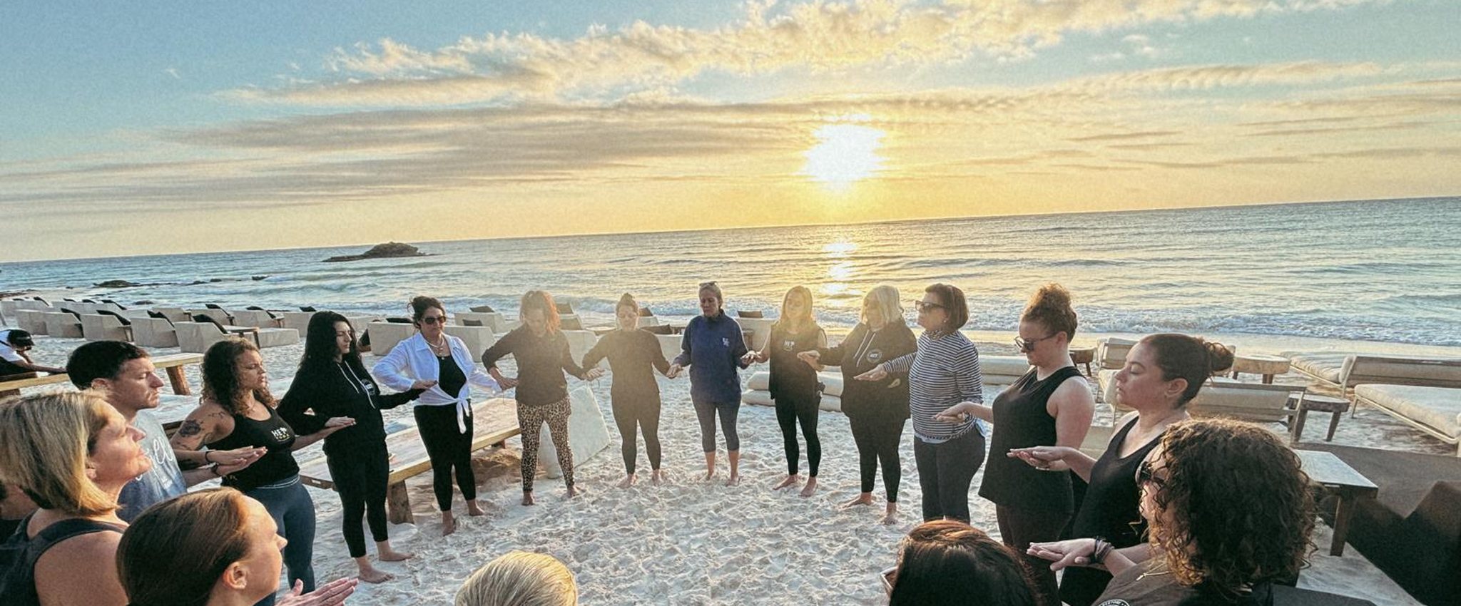 A circle of people holding hands on a beach at sunrise