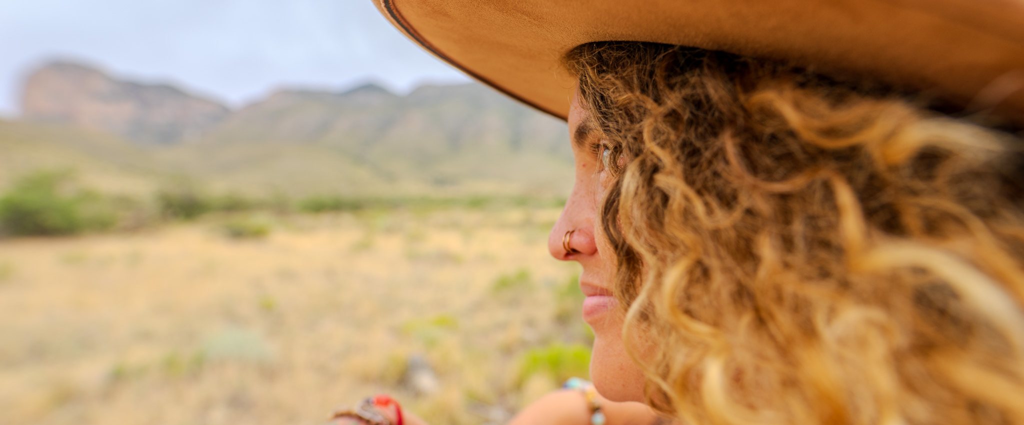 Tasha in profile beneath a wide-brimmed hat, looking out across desert grassland toward distant mountains