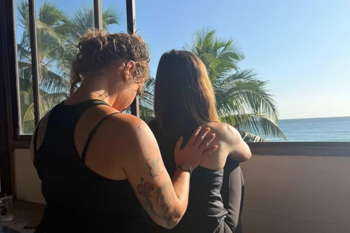 Two retreat guests holding each other by a window looking out at palms and the shore
