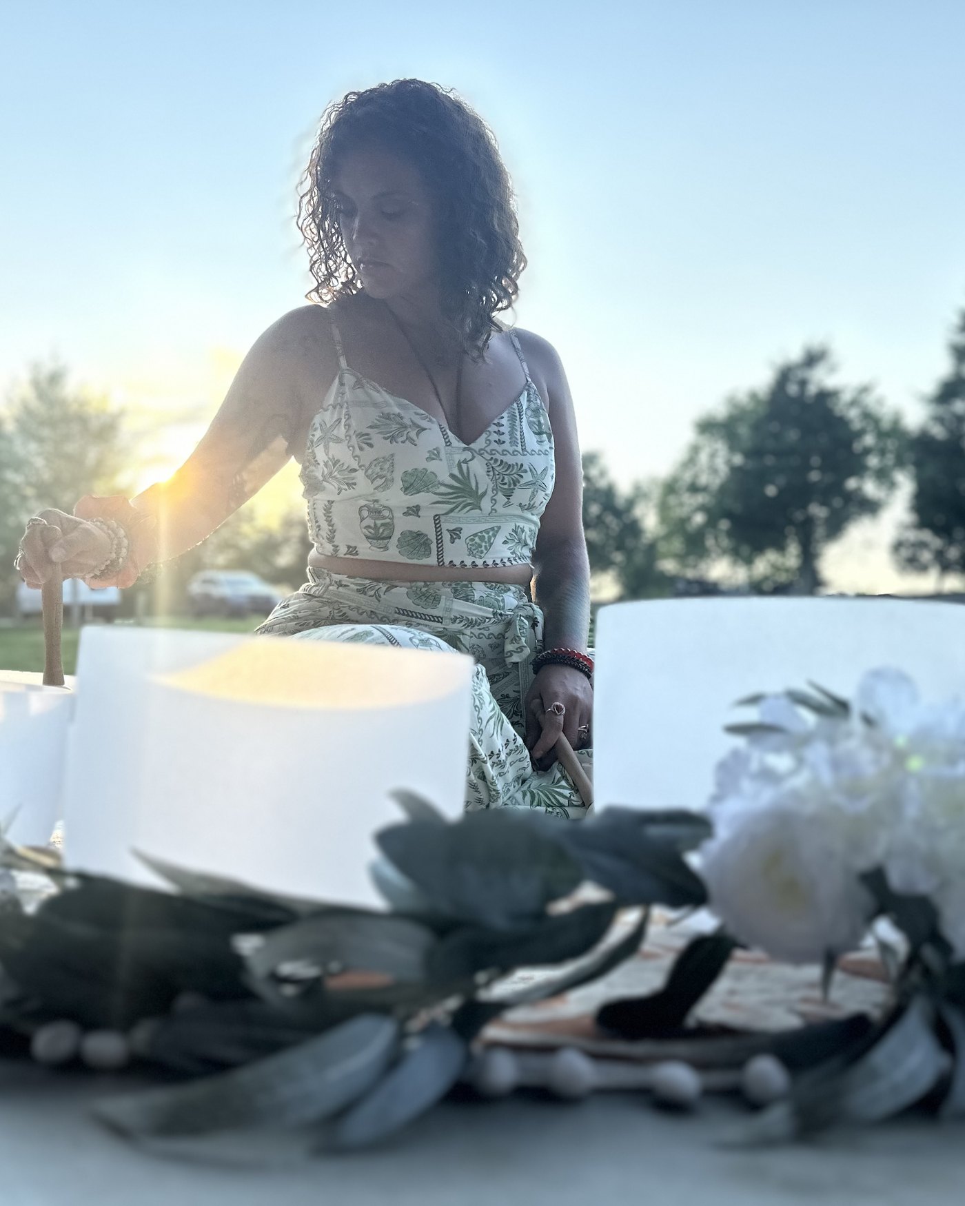 Tasha playing a crystal singing bowl at golden hour, ringed with flowers and greenery